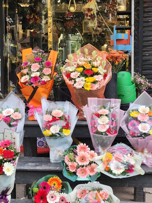 A vibrant display of various floral bouquets arranged on a black shelf outside a flower shop, featuring an assortment of fresh flowers including pink, white, and yellow gerbera daisies, roses, and chr