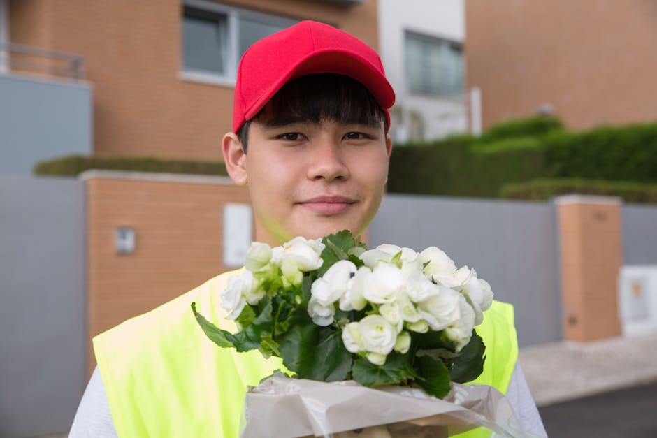 A young man wearing a red cap and a bright yellow high-visibility vest holds a bouquet of fresh white roses wrapped in white paper. The roses are fully bloomed, with smooth, velvety petals, and are co