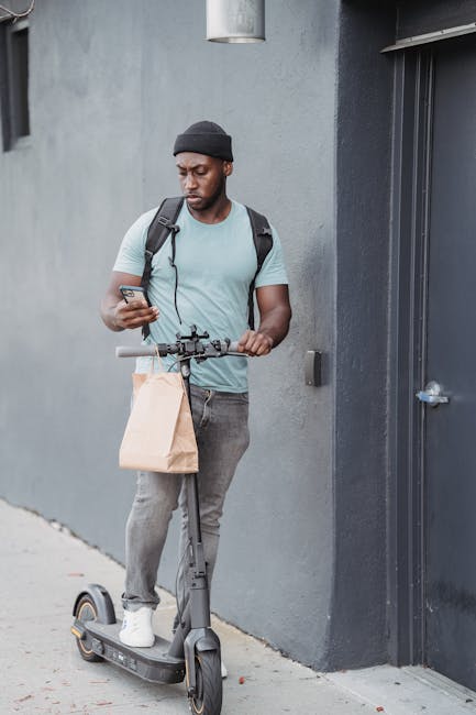 A young man with dark skin, wearing a black beanie, light blue t-shirt, and gray jeans, stands outside next to a gray wall and a door. He is holding a brown paper bag in one hand and is using his smar