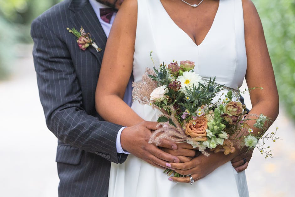 A bride dressed in a white wedding gown is holding a floral bouquet consisting of peach roses, white daisies, green foliage, and delicate pinkish-brown accents. The bouquet is wrapped in natural brown