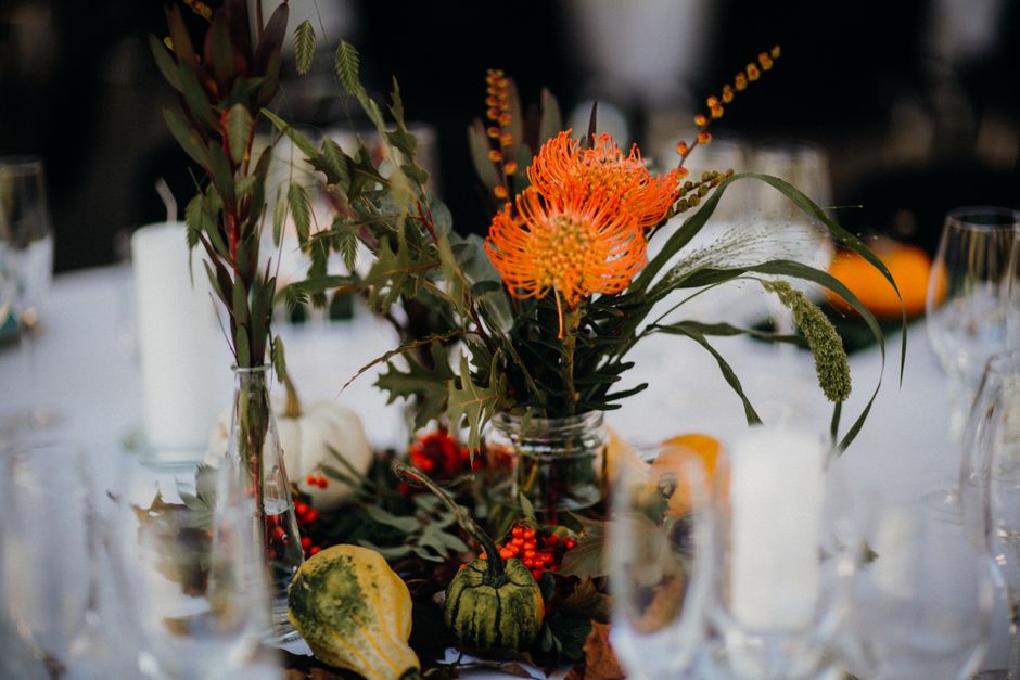A floral arrangement featuring a tall, clear glass vase filled with a mixture of greenery, including spiky, dark green leaves and soft, rounded foliage. Prominently displayed are vibrant orange Protea